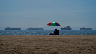 A beach goer sits on the beach in Seal Beach Calif., Friday, Oct. 1, 2021, as container ships waiting to dock at the Ports of Los Angeles and Long Beach are seen in the distance. With three months until Christmas, toy companies are racing to get their toys onto store shelves as they face a severe supply network crunch. Toy makers are feverishly trying to find containers to ship their goods while searching for new alternative routes and ports.