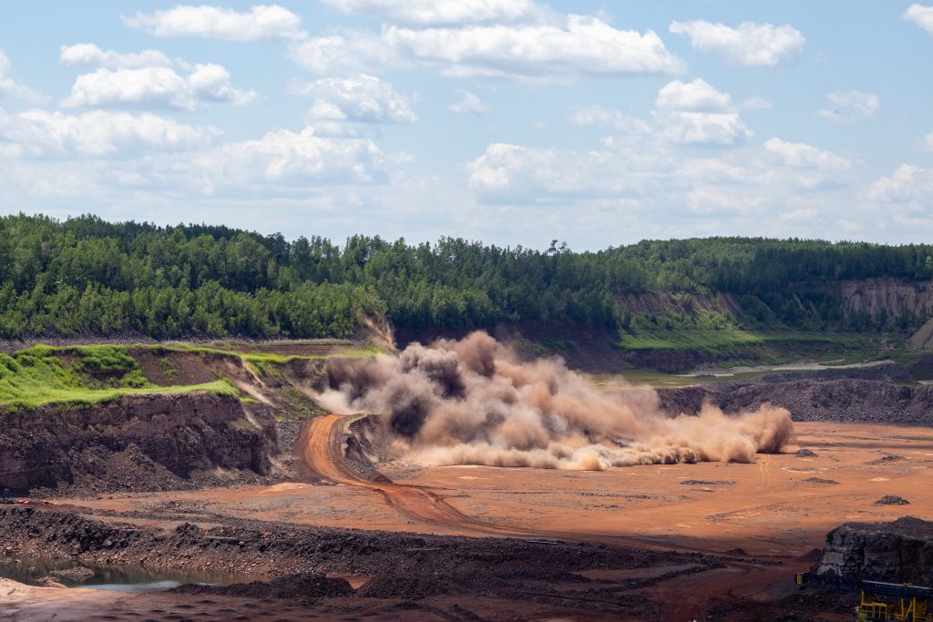 Heavy excavator creating dust cloud while mining in quarry with forested hillside backdrop