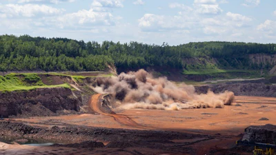 Heavy excavator creating dust cloud while mining in quarry with forested hillside backdrop