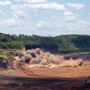 Heavy excavator creating dust cloud while mining in quarry with forested hillside backdrop