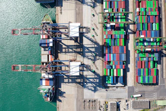 Aerial view of shipping port with cargo cranes, stacked containers, and docked vessel at waterfront terminal