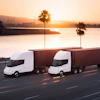 Two white autonomous semi-trucks with cargo containers driving on a highway at sunset with a port and palm trees in the background