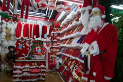Christmas market stall displaying red Santa costumes, white fur trim, and holiday decorations with a young shopper browsing merchandise.