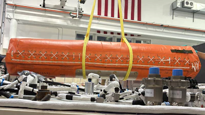 Orange cylindrical aerospace equipment suspended by yellow straps in industrial workshop with American flags