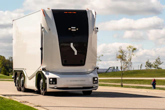 White autonomous semi-truck with black cabin parked on a paved road with green fields and cloudy sky in background