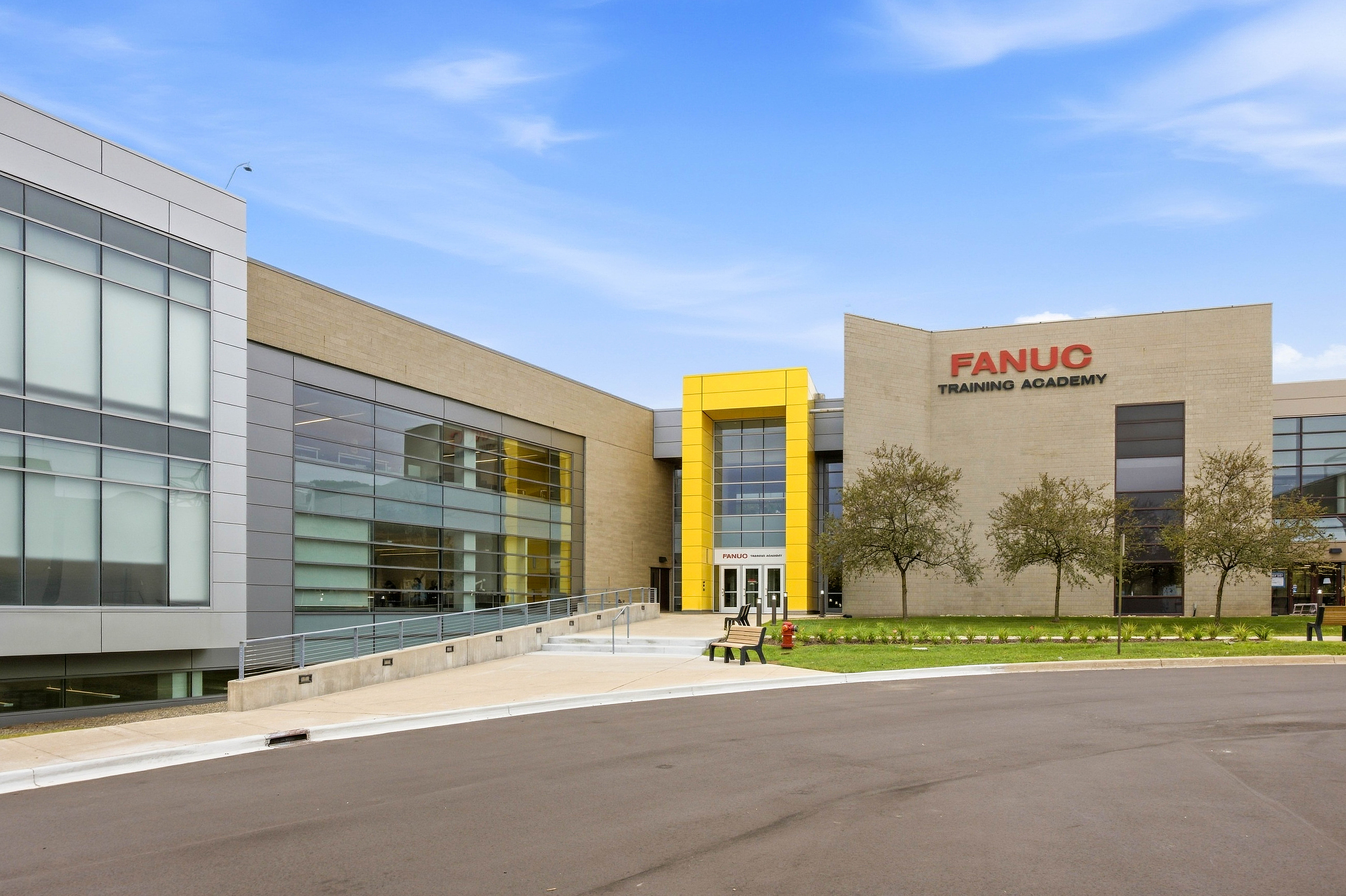 Modern FANUC Training Academy building with glass and tan facade, featuring yellow accent entrance and landscaping