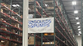 Industrial warehouse interior with tall storage racks, branded signage including Goulds Pumps and Rexnord logos, pallets, and orange safety markers on concrete floor.