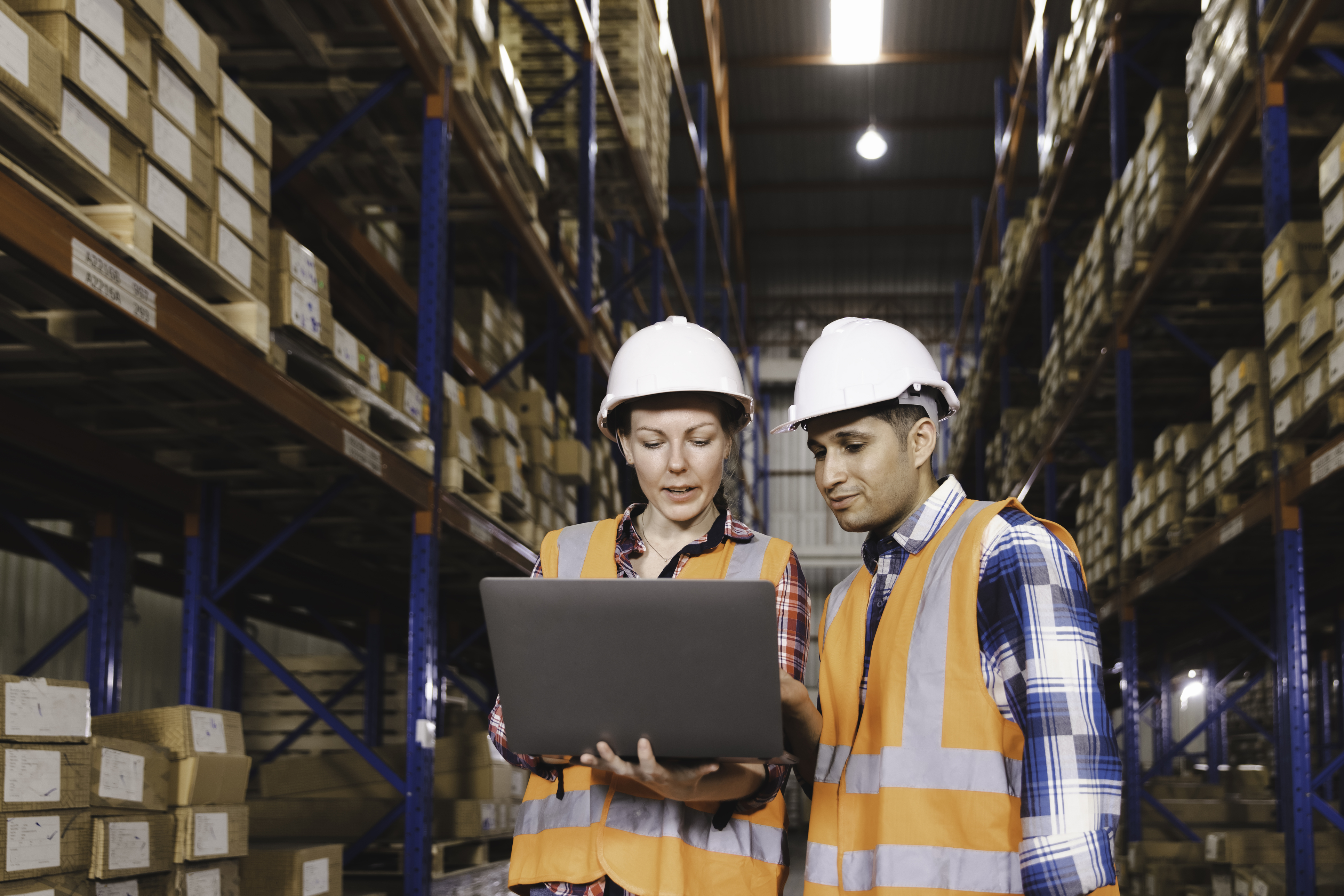 Two warehouse workers in safety gear reviewing inventory data on laptop in storage facility